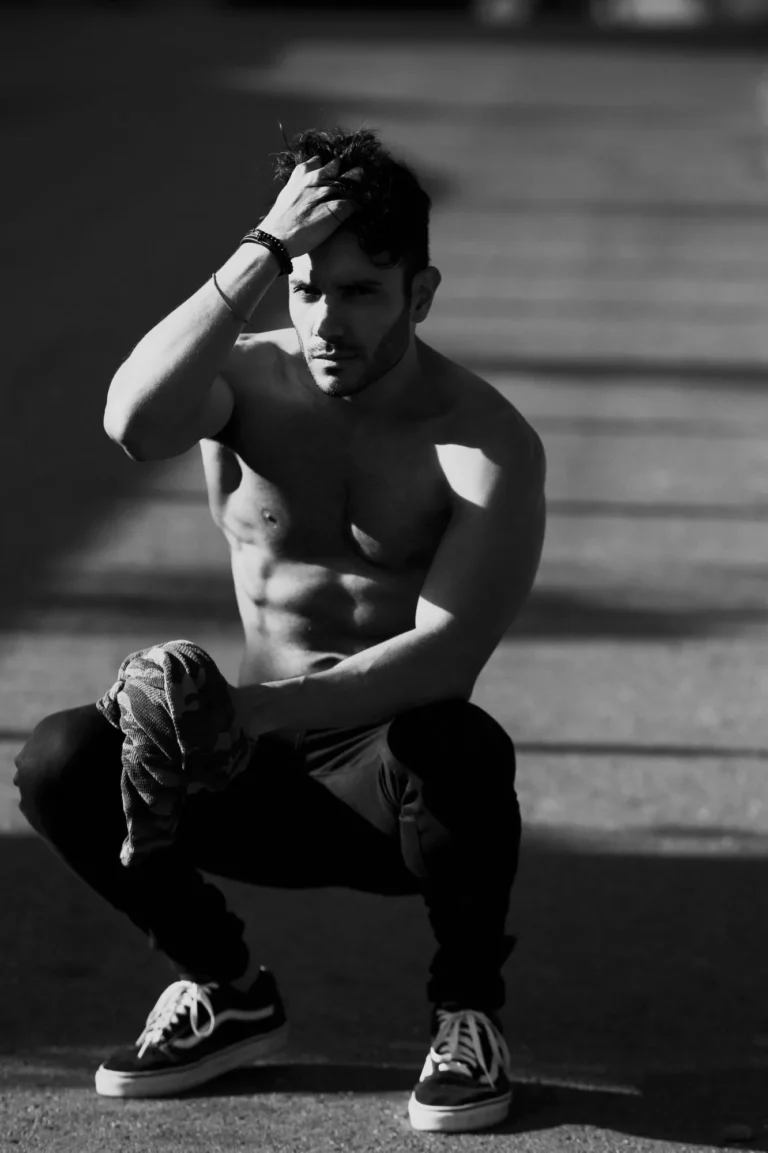 Black and white menswear editorial of a shirtless male model crouching in dramatic sunlight