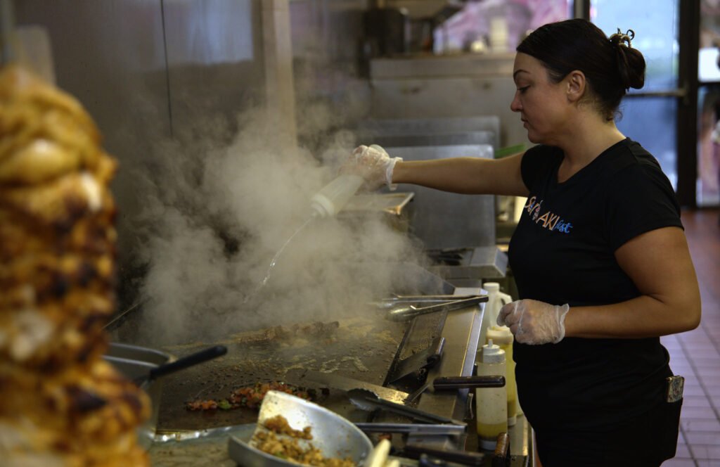 Cook working at a steaming flat-top grill with a gyro spit in the foreground, restaurant kitchen photography