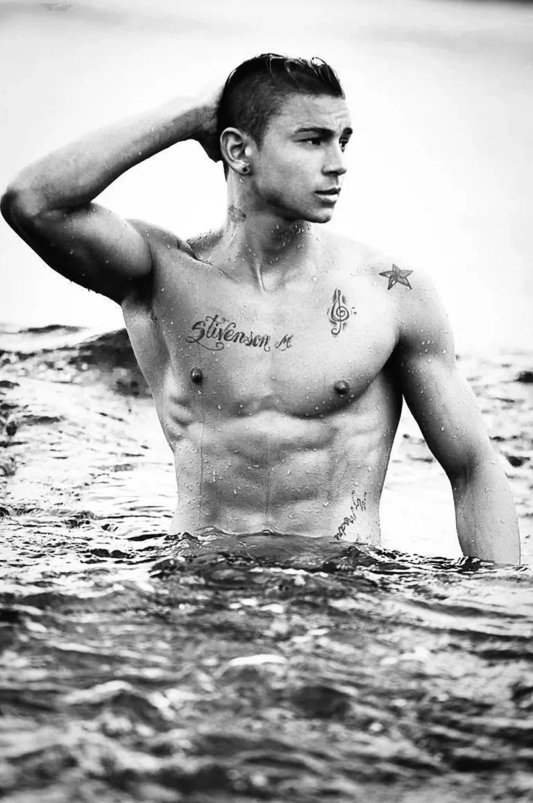 Black and white portrait of a tattooed male model standing waist-deep in the ocean, men's fitness editorial