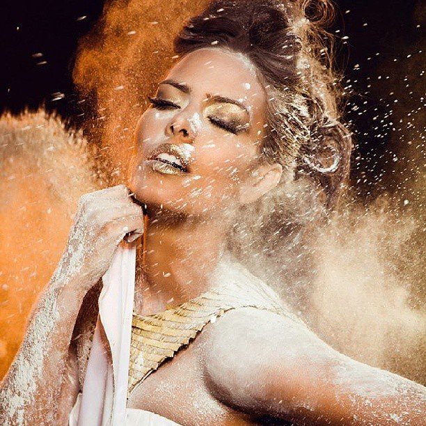 Beauty editorial of a woman with golden makeup surrounded by a burst of white powder against a warm background
