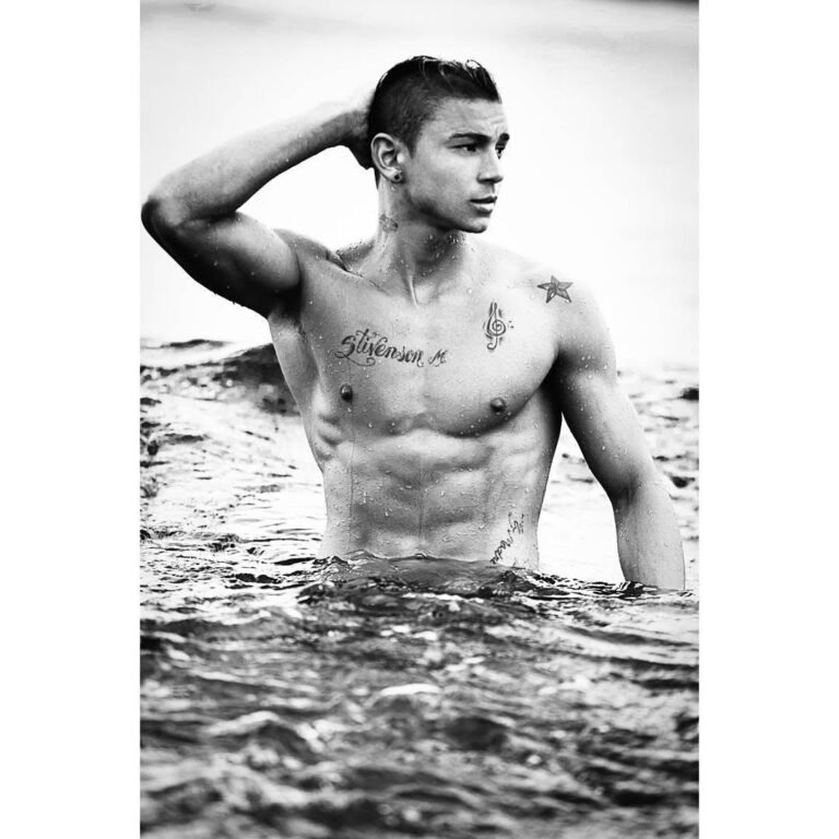 Black and white portrait of a tattooed male model standing waist-deep in the ocean, men's fitness editorial