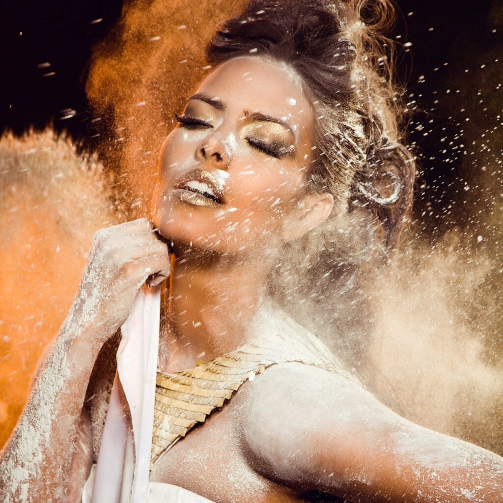 Beauty editorial of a woman with golden makeup surrounded by a burst of white powder against a warm background